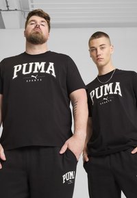 Czarne bawełniane koszulki z białym logo "PUMA SPORTS", zestawione z czarnymi spodniami dresowymi. Oba stroje charakteryzują się luźnym krojem i prostym wzornictwem.