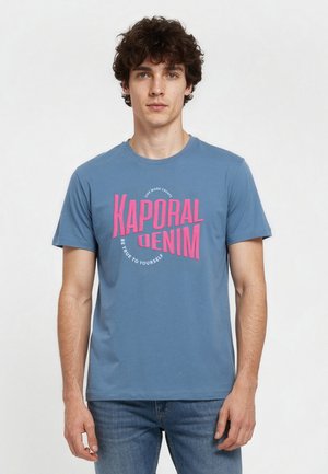 Jeune homme aux cheveux bouclés portant un t-shirt bleu avec un texte rose « Kaporal Denim » et un jean bleu clair, debout devant un fond uni.
