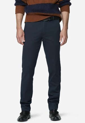 Mand iført mørkeblå bukser, sort bælte, sorte pæne sko og en brun og marineblå stribet sweater, stående mod en hvid baggrund.