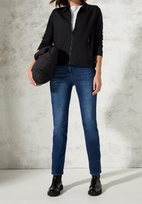 Person in blauen Jeans, schwarzer Zip-Jacke, schwarzen Stiefeln, hält eine schwarze gepolsterte Tasche vor einem hellen strukturierten Wandhintergrund.