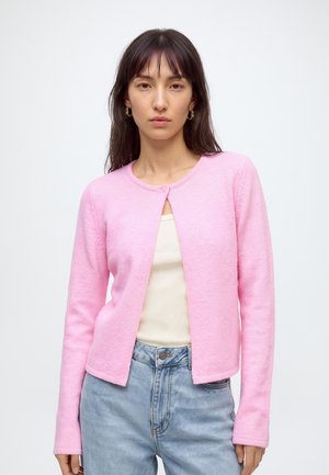 Jeune femme aux longs cheveux foncés porte un cardigan boutonné rose clair sur un haut blanc et un jean bleu clair devant un fond blanc uni.