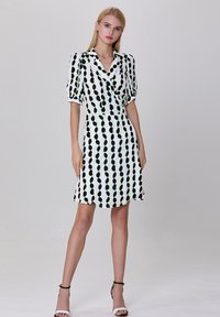 Robe blanche avec des motifs circulaires noirs et verts, manches courtes bouffantes, col en V et ourlet évasé. Portée avec des sandales blanches.
