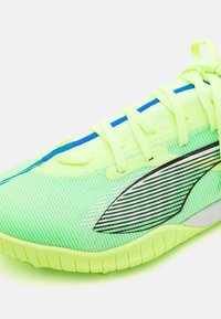 Zapatilla deportiva de color verde neón con parte superior de malla, cordones planos, detalles en azul y una suela texturizada con surcos verticales.