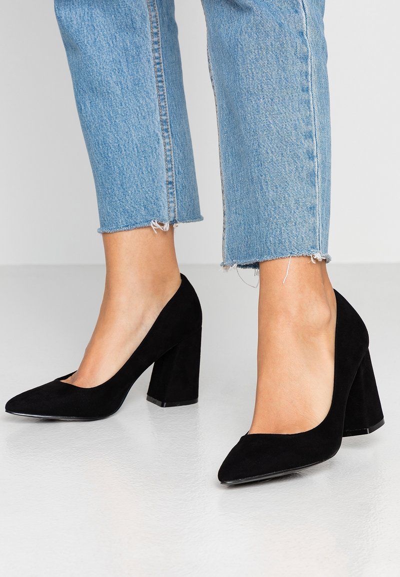 Escarpins noirs en daim avec un bout pointu et un talon épais. Les chaussures ont une texture lisse et sont associées à un pantalon en denim bleu court.