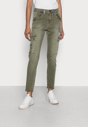 Vrouw draagt slank gesneden olijfgroene cargobroek met rits- en klepzakken, gecombineerd met witte sneakers en een wit shirt.