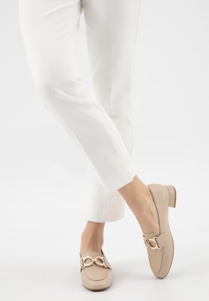 Vrouw draagt witte broek en beige loafers met goudkleurige kettingdetail, staand met één voet over de andere gekruist op een witte achtergrond.