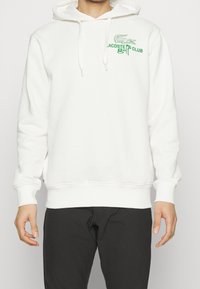 Vit hoodie i mjukt tyg, med en grön Lacoste-logotyp och texten "GOLF CLUB". Den har en känguruficka och en dragsko i huvan.