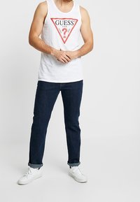 Débardeur blanc sans manches avec un logo triangulaire "GUESS" en rouge et noir, associé à un jean bleu foncé et des baskets blanches. Coupe décontractée.