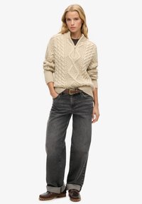 Dikke crème kabelgebreide trui met een halflange ritskraag, gecombineerd met high-waisted, vervaagde zwarte jeans en bruin veterschoenen.