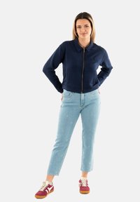 Felpa con zip di colore navy con colletto e logo ricamato, abbinata a jeans azzurri chiari con un motivo sottile e sneakers rosse.