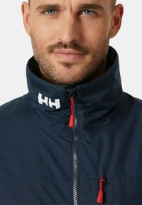 Chaqueta de un navy oscuro con cuello alto, que cuenta con un cierre completo y un tirador de cremallera rojo, acentuada con un logo blanco en el cuello. Textura suave.