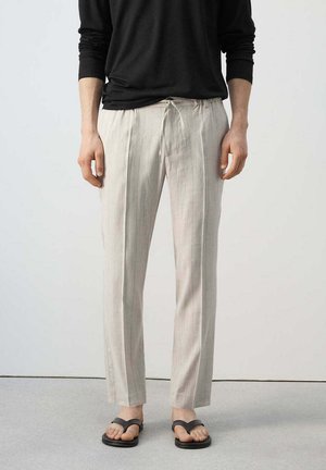 Persona con pantaloni beige a righe sottili con coulisse, top nero a maniche lunghe e sandali neri, in piedi su pavimento grigio.