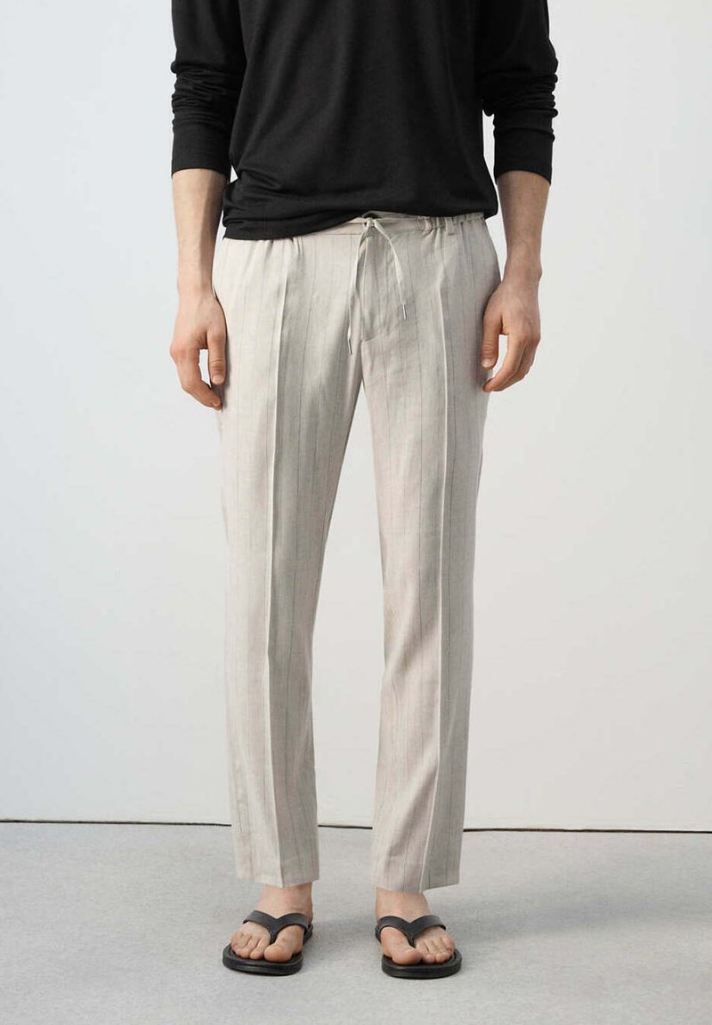 Personne portant un pantalon beige à fines rayures avec cordon, un haut noir à manches longues et des sandales noires, debout sur un sol gris.