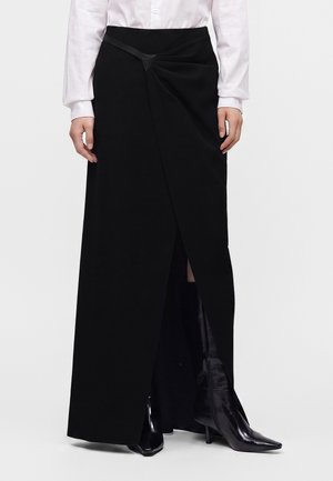 Femme portant une longue jupe portefeuille noire avec une fente à l'avant, associée à des bottes à talons hauts noires à bout pointu et une chemise blanche boutonnée.