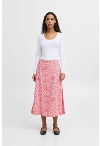 Jupe longue rose avec des motifs en losange, fabriquée en tissu léger. Portée avec un haut ajusté à manches longues blanc et des chaussures plates noires.