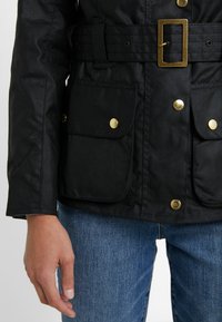 Black jacket with a belted waist, gold hardware, and two front pockets. Features a smooth texture and tailored fit. Worn with blue jeans.