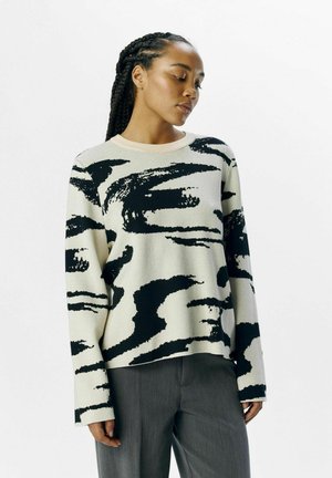 Overdimensioneret sweater i beige med sort abstrakt penselstræk mønster, med rund hals og lange ærmer, lavet af et blødt stof.