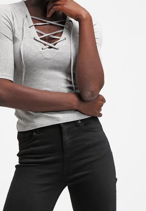 Personne portant un haut gris clair côtelé avec un col lacé et un pantalon noir taille haute, main reposant sur le menton et bras croisés.