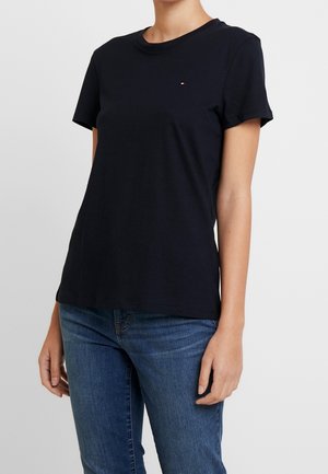 Person trägt ein eng anliegendes schwarzes kurzärmeliges T-Shirt und blaue Jeans und steht vor einem schlichten hellen Hintergrund.