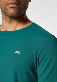 Türkise Langarmshirt aus strukturiertem Material mit einem kleinen, hellgrauen Logo auf der linken Brust und einem Rundhalsausschnitt.