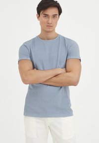 Svetlomodré tričko s krátkym rukávom a okrúhlym výstrihom. Mäkká textúra a minimalistický dizajn. Model má na sebe biele nohavice, prekrížene ruky.