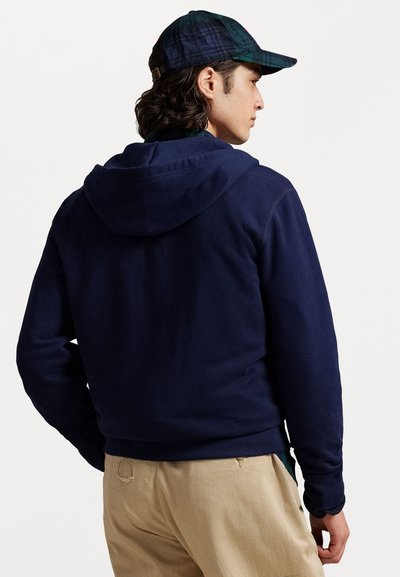 Laivansininen huppari, jossa on sileä pinta. Henkilöllä on päällä vaaleat housut ja siniruutuinen lippalakki.