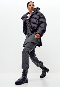 Femeie care merge purtând o jachetă puffer neagră, fustă gri stratificată și pantaloni, cizme negre cu fermoar și cămașă albă cu guler, pe un fundal alb.