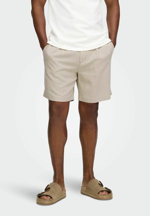 Mann trägt beige Shorts und passende Slip-On-Sandalen, steht mit den Händen in den Taschen vor einem einfarbigen Hintergrund.