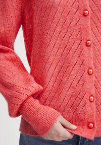 Cardigan lavorato a maglia corallo con un motivo diagonale, dotato di bottoni a forma di perla, polsini a coste e una finitura morbida e texturizzata.