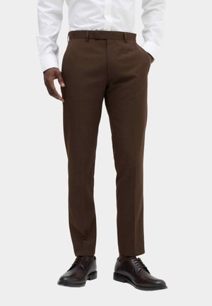 Man wearing brown dress pants and dark brown leather dress shoes with a white long-sleeve shirt, standing with one hand in pocket.