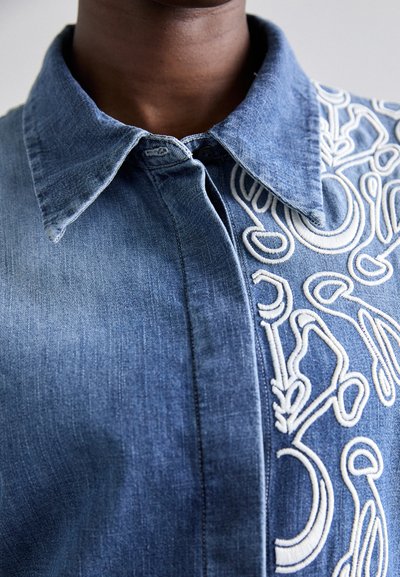 Camicia in denim con colletto, caratterizzata da un ricamo bianco intricato sulla parte anteriore. Il colore è blu medio, con una finitura testurizzata e leggermente sbiadita.