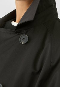 Czarny płaszcz trench z wysokim kołnierzem, gładką teksturą i okrągłym, czterotykowym guzikiem. Tkanina wydaje się lekka i trwała.