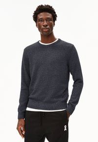 Pull en tricot bleu marine à col rond, en tissu texturé, avec des poignets côtelés. Porté par-dessus un t-shirt blanc, associé à un pantalon de jogging noir avec un accent blanc.