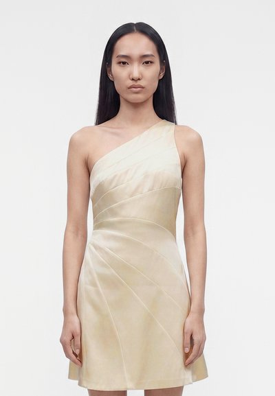 Femme aux longs cheveux noirs portant une robe plissée crème, sans manches, à une épaule, debout devant un fond blanc uni.