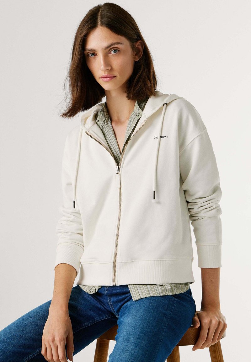 Crèmekleurige hoodie met ritssluiting, trekkoord, geribbelde manchet en zakken, gedragen over een groen gestreept shirt en gecombineerd met een blauwe spijkerbroek.