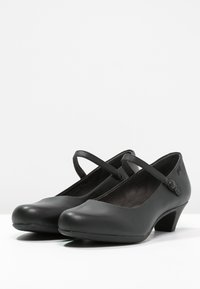 Chaussures Mary Jane en cuir noir avec bout rond, bride ajustable et talon bas. Texture lisse et détail de design subtil près de la bride.