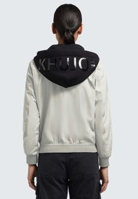 Helehall hall jacket musta kapuutsiga. Omab üle mõõtmete kapuutsi läikiva "KHUJO" tekstiga, ribakäistega ja elastse vöökohtaga.