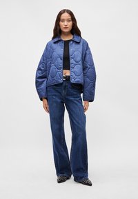 Μπλε καπιτονέ ζακέτα με cropped σχέδιο, μεγάλο γιακά και κουμπιά με πίεση, σε συνδυασμό με φαρδιές τζιν παντελόνες σε σκούρο χρώμα και μαύρα μυτερά παπούτσια.