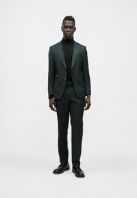 Fato verde escuro feito à medida com lapelas de entalhe, usado sobre uma camisola de gola alta preta, combinado com sapatos pretos brilhantes. Tecido liso, design justo.