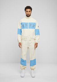 Cream and light blue windbreaker tracksuit with elastic cuffs and waistband. Features "FUBU" branding across the chest. White sneakers complete the look.