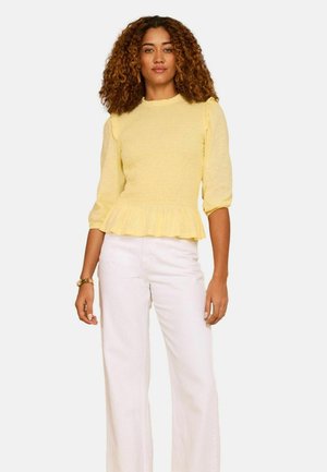 Žena s kudrnatými vlasy, oblečená v žlutém topu s nařaseným lemem a bílých širokých kalhotách s vysokým pasem, stojící před bílým pozadím.