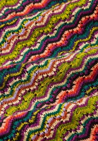 Mehrfarbiger Strickstoff mit einem Zickzackmuster in Grün, Pink, Lila und Creme, mit einer strukturierten Oberfläche und schimmernden Akzenten.