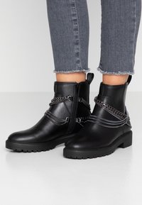 Botas negras de tobillo con un acabado de cuero suave, con detalles de cadenas plateadas y una cremallera lateral, sobre una suela de goma negra texturizada.