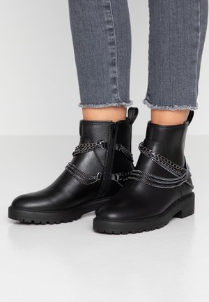 Bottines noires avec une finition en cuir lisse, ornées d'accents en chaîne argentée et d'une fermeture éclair latérale, sur une semelle en caoutchouc noir texturée.