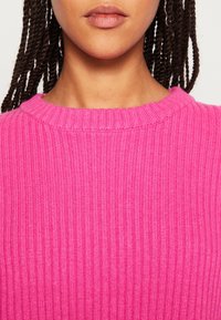 Gros plan d'une personne aux cheveux tressés portant un pull côtelé rose vif, montrant le cou, le bas du visage et le haut du torse.