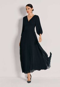 Zwarte geplooide jurk met een V-hals en lange mouwen. Gemaakt van lichtgewicht stof, valt tot aan de enkels met een vloeiende silhouet.