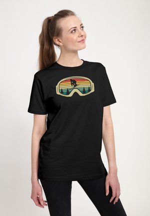 Kvinde står og smiler iført en sort t-shirt med en skibrille-grafik, der viser en snowboarder, træer og en solnedgang inde i linsen.
