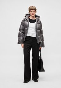 Pihepuha szürke kapucnis dzseki, fényes felülettel, cipzáras záródással, fekete felső és fekete nadrág fölött viselve, fekete bevásárlótáskával párosítva.