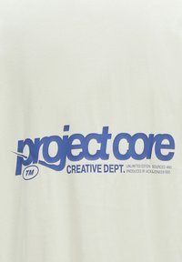 Hvid bomulds t-shirt med en stor blå grafisk tekst, der står "project core CREATIVE DEPT." med yderligere tekst nedenfor.
