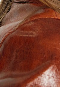 Brun läderaxelväska med en slät, glänsande yta. Synlig sömnad längs sömmarna och en antydan av hår på toppen.